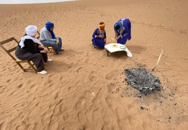 Guides in blue robes preparing a traditional meal for two travellers in the dunes