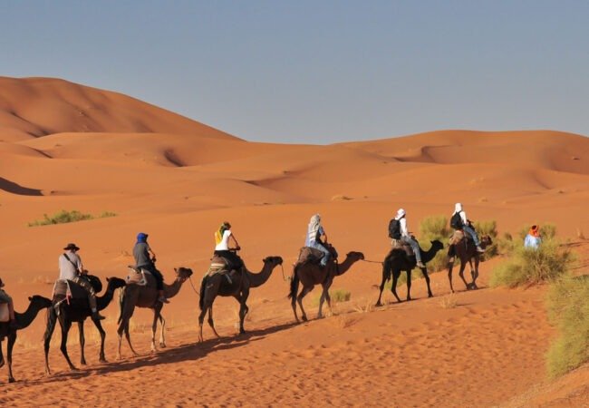 Riders on camels crossing orange dunes with a guide walking beside them